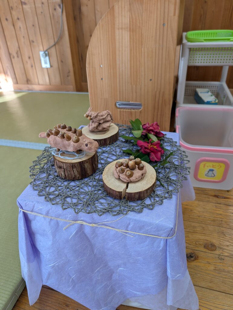 薄紫色の布を被せた台の上に、切り株の土台に乗った粘土の作品が3つ並んでいる。粘土にはドングリが埋め込まれ、生き物のような形をしている。