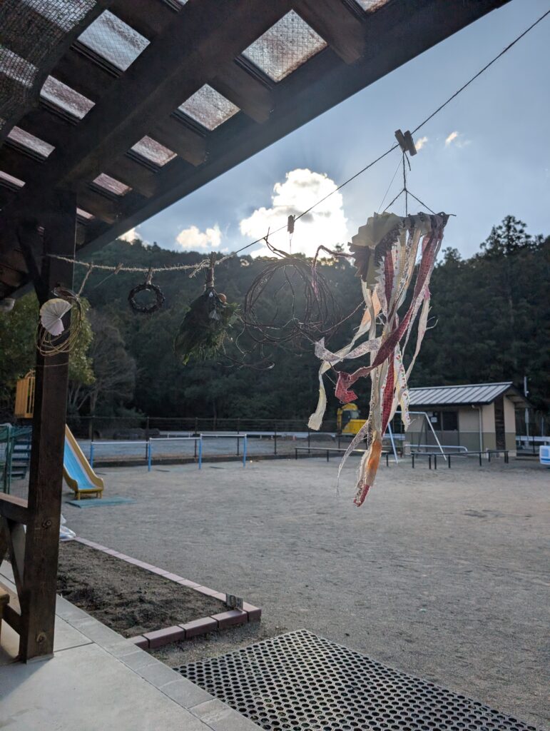 園庭を見渡すテラスの軒下。蔓（つる）を丸めて細長く裂いた布切れをつけた帽子やオーナメンのようなものが逆光の中で風に揺れている。