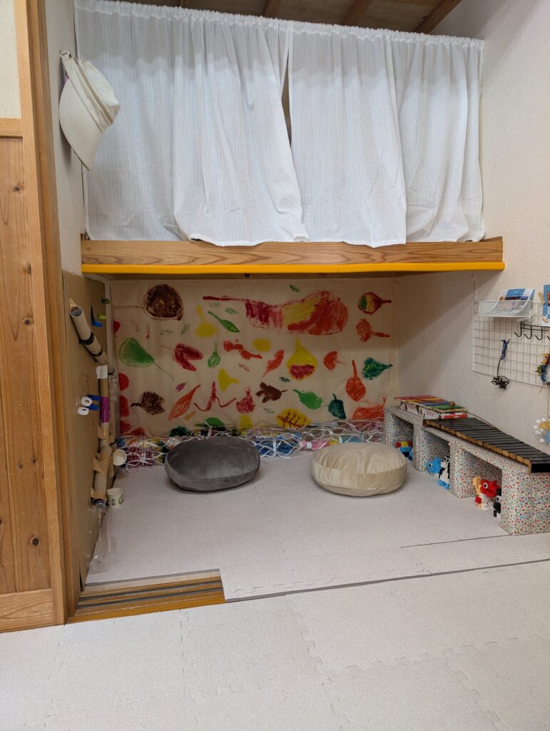 押し入れの下段を活用した「秘密基地」のような空間。床にはマットが敷かれ、丸いクッションが2つ置かれている。奥の壁には色鮮やかな絵が飾られ、手前には手作りの楽器（木琴など）が並ぶ。
