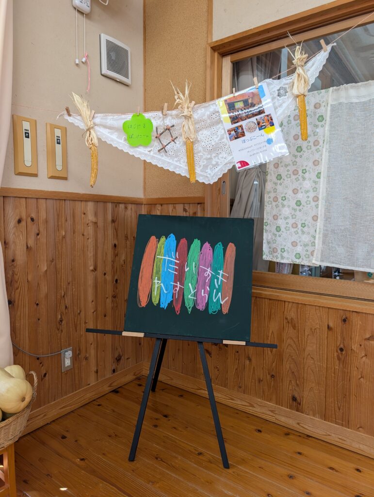 「だいすきれすとらん」と書かれた手描きの黒板看板。背後の紐には、トウモロコシや活動の様子を伝える写真がディスプレイされている。