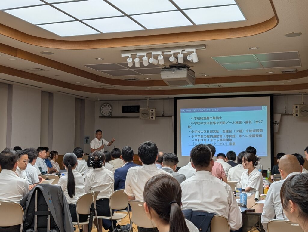 手前にはスーツ姿の参加者たちの後ろ姿、奥に森四日市市長がマイクを持って立っている。スクリーンにはスライドが投影されている「小学校給食の無償化・小学校の水泳指導を民間プール施設へ委託（37校）・中学校の休日部活動　全種目(16種)を地域展開・小中学校の屋内運動場(体育館)等への空調整備→令和9年4月から供用開始・こどもみらいクーポン事業
